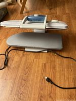 Photo showing electric hand press steamer iron set on wooden floor, with grey fabric covered ironing board and electric cord visible.