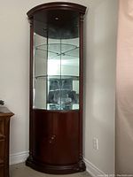 Full view of the dark brown mahogany corner curio cabinet showing curved glass front, three glass shelves, mirrored back, and light at the top. Lower storage doors visible.