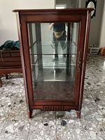 Front view of a mahogany display cabinet with glass door and brass handle, showing two internal glass shelves and reflecting room interior.