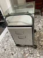 Front view showing glass top, two drawers with silver metal pull handles, and wheels.