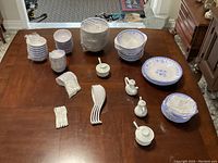 Photo showing various stacks of rice bowls, soup bowls wrapped in plastic, plates with blue floral pattern, white melamine spoons arranged on a table, small packets of small spoons, and two small white containers.