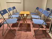 Front angle showing 6 blue folding chairs arranged in a semicircle around a small foldable wooden table on a carpeted floor.
