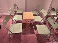 Photo of six light green plastic folding chairs arranged around a small rectangular wood folding table on a red carpeted floor in a room with mirrored wall