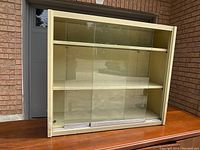 A front view of the ivory steel cabinet showing both sliding glass doors closed, two internal shelves visible, placed on a table outside a garage.