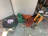 Wide shot showing all items together on patio ground: two coiled black sprinkler hoses, several coiled orange and black extension cords, green plastic watering can.