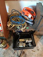 Multiple extension cords wound and loosely coiled, an orange cord winder, and some other electrical items on a wooden surface and in a crate.