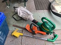 Photo of three garden tools including Weed Eater blower vac, Weed Eater trimmer, Black & Decker hedge trimmer on floor with dust bag attached to blower.