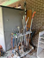 Photo shows gardening tools including rakes, shovel, pruners, and handsaw stored upright in a beige plastic garden tool rack near a brick wall and storage cabinet.
