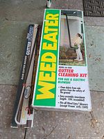 Photo of Weed Eater GA-2000 gutter cleaning kit box with prominent green and yellow branding and usage instructions, next to Supertek Rain Gutter Flusher in black packaging.