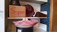 Shelf with a large colorful Mexican sombrero and a brown suede cowboy hat with feather decoration. Box labeled 'Simpson's' under dark straw fedora.