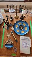 Photo showing assorted Judaica items on table: brass Menorah, blue Seder plate, candle holders, decorative Star of David, trivet, keychain, and figurine.