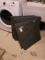 Stack of black square interlocking foam floor mats leaning against a washing machine in a laundry room.