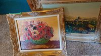 Two large framed paintings rest on carpet, left floral painting showing a colorful floral bouquet in green vase with gold frame, right painting showing cityscape of Jerusalem with ornate gold frame.