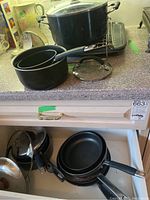 Pots and pans displayed on kitchen counter and drawer, showing various pots with lids, frying pans, and a pie plate