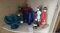 Blue 6-inch bowl on pedestal, vintage green vase with gold trim, red 12-inch bowl with lid on stand, two signed Willy Woodward figurines, three colored glass vases about 7 inches tall on a corner shelf