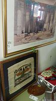 Framed large print showing columns and hieroglyphics of Philae Temple with a gold frame, papyrus-style framed print with Egyptian motif in black frame below, and oval wood art on shelf.