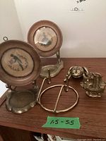 Photo showing two brass stands holding framed embroidered pictures of a man and a woman, along with brass frog figurine, ship figurine, and plate holder on wooden surface.