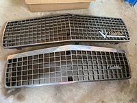 Top view of two Cadillac front grilles laying on a concrete floor, one with V8 emblem and Cadillac script badge on the right side, the other without an emblem.