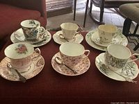 Front view of all 5 teacup, saucer, and plate sets arranged on red surface, showcasing floral and botanical designs, alongside four collectors teaspoons.