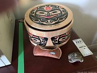 Top view of round wood box showing painted stone face Indigenous design and signature on lid, along with small stone face piece and canoe-shaped item on side