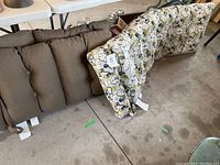 Photo showing one brown lounge cushion with tufted buttons alongside two floral print outdoor chair cushions, all placed on a concrete floor.