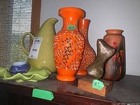 Green Russel Wright style pitcher, orange ceramic cutout vases, pottery vase with floral design, metal abstract sculpture on wooden base, small bowls and trays on a wood surface near window blinds.