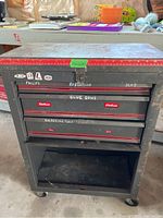 Closed black metal wheeled tool box with label on drawers indicating contents like glue guns and soldering tools. Shows surface rust and wear.