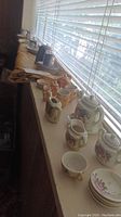 Wide view of assorted miniature tea sets and ceramics arrayed on a window sill, showing multiple tea set pieces in various colors and styles.