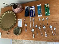 Full lot overview showing large brass tray, copper kettle, round brass mesh container, and assorted spoons and forks arrayed on a wood table.