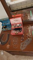 Overview of assortment of costume jewelry laid out on dresser with two jewelry boxes (one blue, one pink) open showing smaller jewelry items inside.