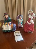 Three porcelain figurines displayed on a wooden surface, including a seated older woman, a young girl with an umbrella, and a woman in red dress with white cloak.