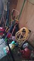 Assorted garden tools grouped including rakes, shovels, extension cord, and watering can stored indoors