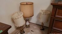 Photo showing three table lamps on floor: one porcelain-based with taller beige shade, two metal-based with ornate bases and fabric shades with floral ribbon trim