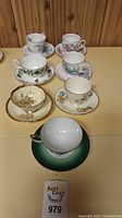 All seven tea cups and matching saucers arranged on table showing variety of floral patterns and gold trim.