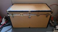 Full view of the rectangular vintage trunk chest showing tan panels with black reinforced edges and metal hardware locks.