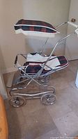 Vintage baby pram with plaid fabric and white liner seen from the side, showing metal frame and four wheels.