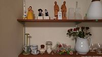 Shelf with wood carved figurines, ceramic pitcher, artificial flowers, and assorted ceramics.