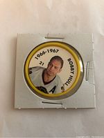 Front side of the coin shows a color photo of Bobby Hull with text '1966-1967, Bobby Hull 21' on a yellow background, held in a square white cardboard holder with staples.
