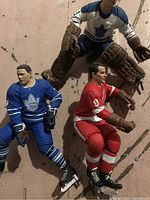 Three loose NHL action figures shown on a brown surface. One goalie in blue and white Toronto Maple Leafs uniform with brown pads. Two skaters, one in blue Maple Leafs uniform, one in red Detroit Red Wings uniform.
