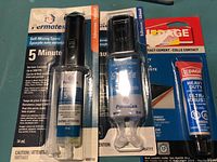 Photo of two Permatex 5 Minute Epoxy kits (14 ml and 25 ml) and one Lepage Contact Cement tube in packaging on a teal surface.
