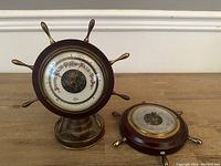 Both barometers displayed together on a wooden surface, showing the brass pedestal version upright and the wall-mounted version flat.