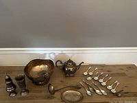 Full view of the silverplate lot including bowl, teapot, salt shaker, ladle, spoons, cups, and small tray on wooden table