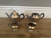 Four item set of silverplate tableware on wooden surface: two teapots (one with lid, one without), creamer, sugar bowl.