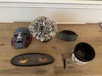 Photo showing six decorative pieces on a wooden surface including the mosaic mask, floral arrangement, metal piece, ceramic pot, metal tray with fish, and keychain.