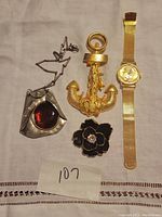 Full view of four items: abstract silver-tone pendant with amber center, gold-tone anchor brooch, black flower pendant, and gold-tone wristwatch