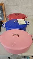 Three oval enameled cast iron Dutch ovens in pink, blue, and red with lids on a table.