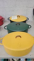Three enameled cast iron Dutch ovens in yellow and green with lids and gold handles, displayed side by side showing exterior colors and condition