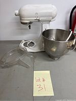 White KitchenAid stand mixer with attached power cord and stainless steel mixing bowl containing whisk and dough hook attachments, plus a paddle attachment resting inside the bowl. Clear plastic pouring shield placed beside mixer.