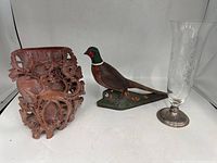 Front view of all three items: reddish-brown carved ceramic vase on left, wooden pheasant bird sculpture center, tall clear glass vase with engravings and metal base on right.