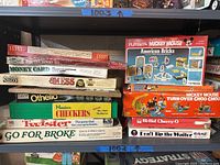 Shelf view of multiple boxed vintage board games mainly stacked and arranged, showing clearly the titles and brands including Playskool Mickey Mouse American Bricks, Milton Bradley Twister, Hasbro Checkers, and others.